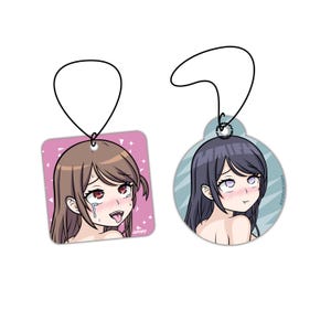 May include: Two car air freshener charms featuring anime characters. The first charm is square and pink with a girl with brown hair and red eyes. The second charm is round and blue with a girl with dark blue hair and blue eyes. Both charms have a string for hanging.