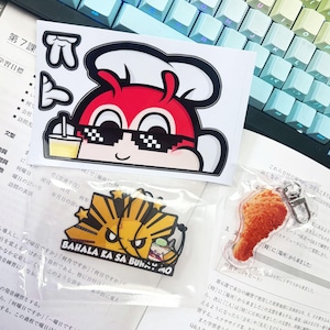 May include: A collection of stickers and keychains featuring cartoon characters. One sticker shows a red character with sunglasses and a chef's hat. A keychain depicts a fried chicken leg. Another keychain has a yellow and black design with text.