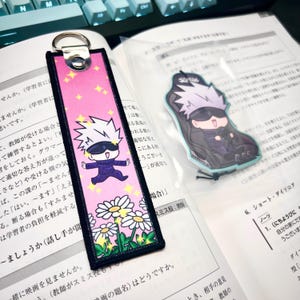 May include: A pink bookmark with a cartoon character and daisies. The bookmark has a silver ring at the top. A clear plastic bag contains a character-shaped charm. The background includes a keyboard and open book.