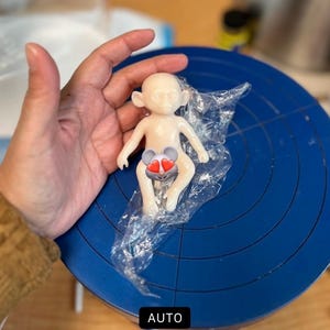 May include: A small, off-white figurine of a monkey-like creature with large ears, sitting on a piece of plastic wrap. The figurine has a heart-shaped design on its torso. The figurine is on a blue surface.