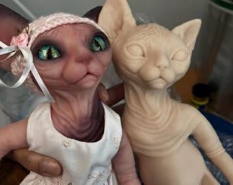 May include: Two Sphynx cat dolls. One doll is wearing a pink dress and a matching hat with a flower. The other doll is nude. The doll on the left has large, detailed green eyes. The dolls are held by a person.