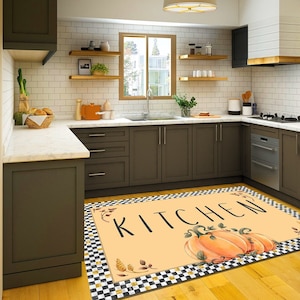 May include: A kitchen rug with a light orange background featuring the word "KITCHEN" in black script. The rug has a black and white checkered border and illustrations of pumpkins and fall foliage.