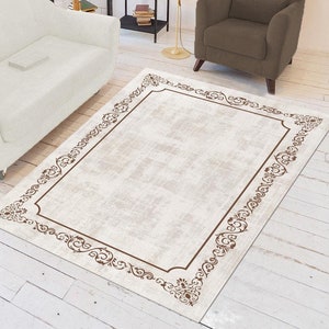 Puede incluir: Una alfombra beige con un diseño de borde ornamentado marrón. La alfombra está en un salón con un sofá blanco y un sillón marrón.