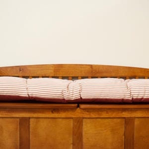 May include: A wooden bench with a red and white striped cushion. The bench is made of stained wood and has a backrest and armrests. The cushion is rectangular and has a classic striped pattern.