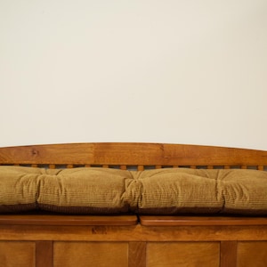 May include: Wooden bench with a tufted, corduroy-like cushion in shades of brown and gold. The bench has a slatted back and armrests, and is made of stained wood. The cushion is divided into three sections.