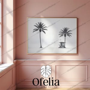May include: A black and white photograph of two palm trees and a lifeguard stand on a beach. The photograph is framed in a gold frame and hanging on a pink wall. The text "Ofelia INTERIOR DESIGN" is below the frame.