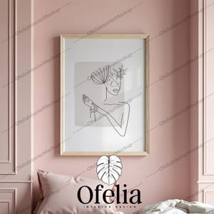 Può includere: Una stampa artistica incorniciata con un disegno a tratto minimalista del viso e del corpo di una donna, con elementi floreali e geometrici. L'opera d'arte è ambientata su uno sfondo grigio chiaro all'interno di una cornice di legno. L'immagine è esposta su una parete rosa, con un cuscino e biancheria da letto sottostanti. Il marchio "Ofelia" è in basso.