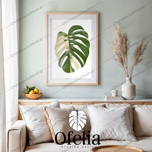 Può includere: Una stampa incorniciata di una grande foglia di monstera variegata verde e bianca su sfondo bianco. La cornice è in legno chiaro ed è appesa a una parete verde chiaro. La stampa è in un soggiorno con un divano, cuscini e un vaso di erba della pampa secca.