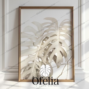 Può includere: Una stampa incorniciata di una foglia di monstera bianca con una cornice dorata. La stampa ha il testo "Ofelia Interior Design" in basso.