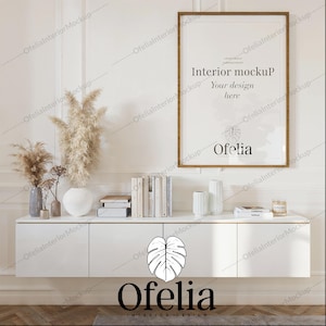 Può includere: Un armadio bianco con due ante e un quadro incorniciato sopra. La cornice del quadro è in legno marrone e l'immagine è bianca con il testo "Interior mockup Your design here" e la parola "Ofelia". Ci sono erba della pampa secca e altre piante in vasi sull'armadio.