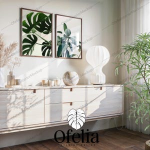 Può includere: Due stampe botaniche incorniciate con cornici in legno scuro sono appese sopra un mobile in legno chiaro. Una lampada bianca e un vaso decorativo sono sul mobile, insieme a candele. Una grande pianta verde è nell'angolo. Il logo Ofelia Interior Design è in basso.