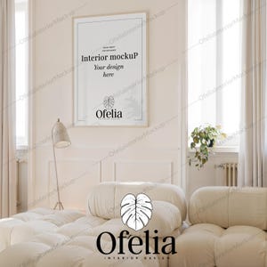 Può includere: Un divano bianco con due cuscini in un soggiorno con un poster incorniciato che dice "Interior mockup Your design here Ofelia" e una pianta in vaso nell'angolo.