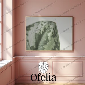 May include: A framed print of a close-up of a green leaf with a white background. The frame is gold and the print is hanging on a pink wall. The text "Ofelia Interior Design" is below the print.