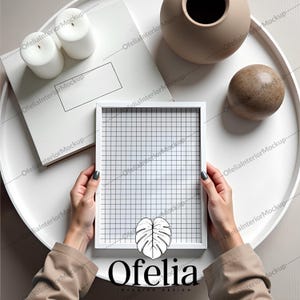 May include: A white picture frame with a grid pattern inside the frame. The frame is being held by a person's hands. The frame is on a white table with a white book, two white candles, a brown vase, and a brown ball.