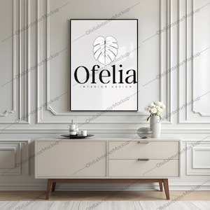 Può includere: Una stampa artistica incorniciata con un'illustrazione in bianco e nero di una foglia di monstera e la scritta "Ofelia INTERIOR DESIGN". La stampa è sopra un mobile color crema con gambe in legno, un vaso di fiori bianchi e oggetti decorativi. Il mobile è largo circa 150 cm.