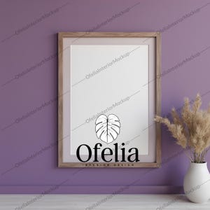 May include: A framed print with a white background and a black and white illustration of a leaf. The text "Ofelia INTERIOR DESIGN" is printed below the leaf. The frame is brown wood and is hanging on a purple wall. A vase with dried pampas grass is on a wooden floor in front of the frame.