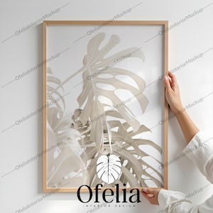 May include: A framed print of a white monstera leaf with a light brown wooden frame. The print is hanging on a white wall. The text "Ofelia Interior Design" is at the bottom of the print.