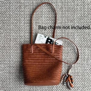 Peut inclure: Un sac fourre-tout marron tiss&eacute; avec une longue anse et une anse plus courte. Le sac contient un magazine. Un charm d&eacute;coratif est attach&eacute; &agrave; la poign&eacute;e la plus courte. Le texte sur l'image indique : "Bag charm not included."
