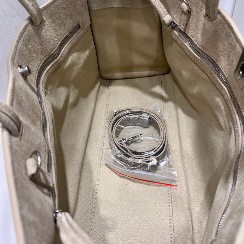 May include: Beige suede tote bag with a matching shoulder strap. The interior of the bag is visible, showing a zippered pocket and a light-colored lining. The strap is packaged in a clear plastic bag.