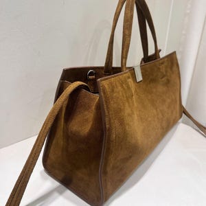 May include: A brown suede tote bag with a rectangular shape. The bag has two top handles and a long, detachable shoulder strap. A silver-colored metal bar secures the top. The bag's interior is visible.