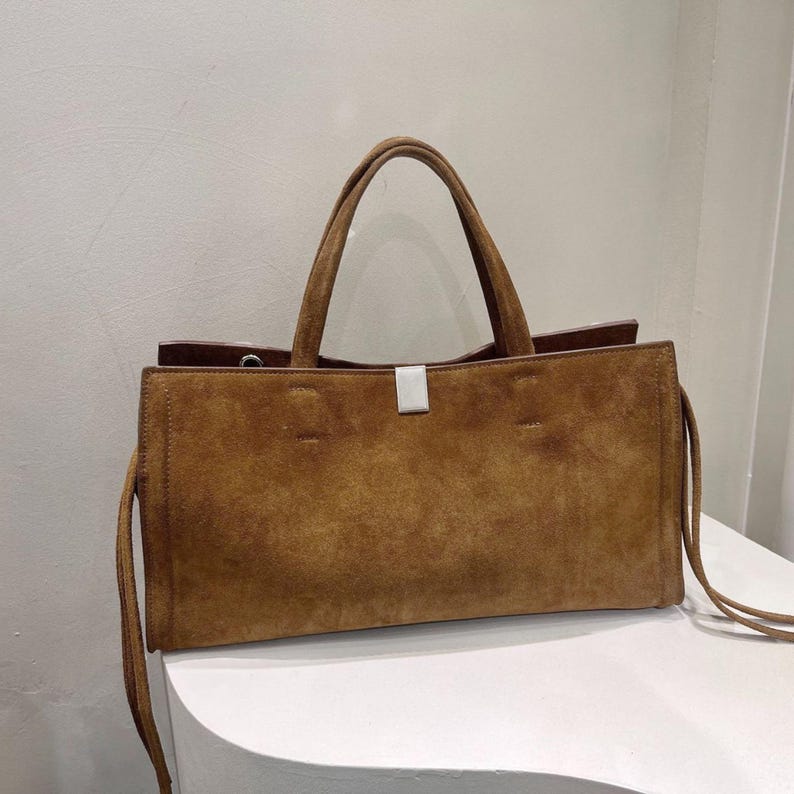 May include: A brown suede tote bag with two top handles and a rectangular shape. The bag features a silver-colored clasp and long, thin straps on the sides. The bag is sitting on a white surface.