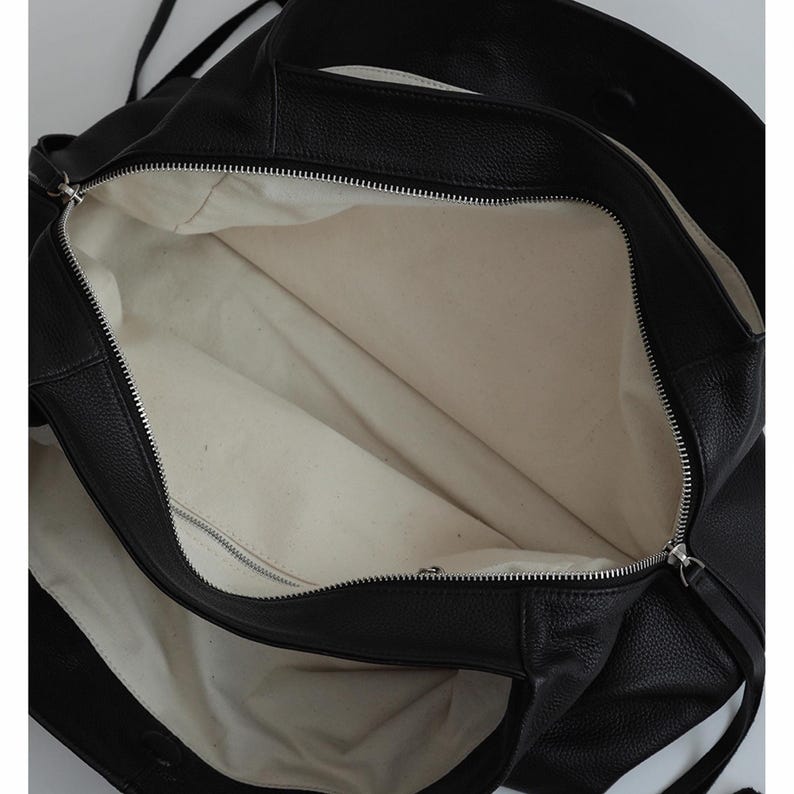 K&ouml;nnte beinhalten: Schwarze Ledertasche mit silbernem Rei&szlig;verschluss. Der Innenraum ist mit einem hellbeigen Stoff gef&uuml;ttert. Die Tasche hat lange Tr&auml;ger zum &Uuml;ber-der-Schulter-Tragen.