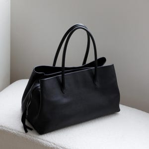 K&ouml;nnte beinhalten: Eine gro&szlig;e, schwarze Ledertasche mit zwei oberen Griffen. Die Tasche hat eine rechteckige Form mit einem Rei&szlig;verschluss an der Seite. Die Tasche liegt auf einer wei&szlig;en, strukturierten Oberfl&auml;che.