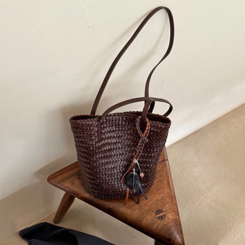 Peut inclure: Un sac fourre-tout tiss&eacute; brun fonc&eacute; avec de longues et fines anses. Le sac pr&eacute;sente un motif tiss&eacute; et une breloque d&eacute;corative avec un charme rond noir et de petits accents de cuir. Le sac est pos&eacute; sur un tabouret en bois.
