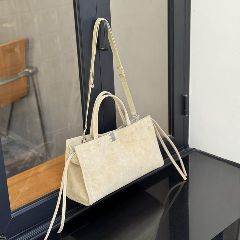 May include: A beige suede tote bag with a shoulder strap and two top handles. The bag has a rectangular shape with a silver-colored metal clasp. Long, thin straps hang from the sides of the bag. The bag is displayed near a window.