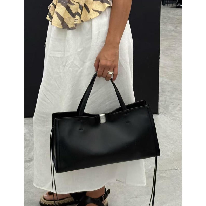 May include: A black rectangular handbag with two handles and a silver accent. The bag is held by a person wearing a white skirt and a top with a yellow and brown pattern. The bag has a minimalist design.