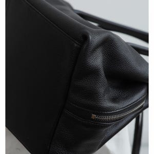 K&ouml;nnte beinhalten: Nahaufnahme einer schwarzen Ledertasche. Die Tasche hat eine strukturierte Oberfl&auml;che und einen silbernen Rei&szlig;verschluss. Das Design der Tasche ist schlicht und elegant, f&uuml;r den t&auml;glichen Gebrauch geeignet.