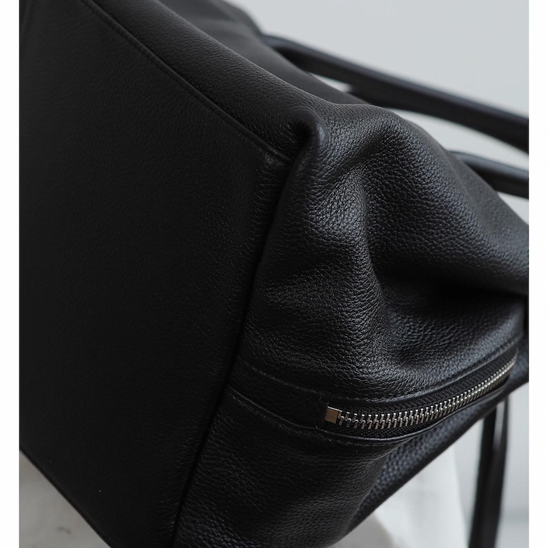 K&ouml;nnte beinhalten: Nahaufnahme einer schwarzen Ledertasche. Die Tasche hat eine strukturierte Oberfl&auml;che und einen silbernen Rei&szlig;verschluss. Das Design der Tasche ist schlicht und elegant, f&uuml;r den t&auml;glichen Gebrauch geeignet.