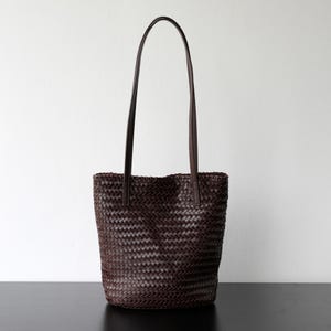 Peut inclure: Un sac fourre-tout en cuir tress&eacute; marron fonc&eacute; avec de longues et fines anses. Le sac a une forme rectangulaire avec un motif tiss&eacute; sur tout le corps. Les anses sont fix&eacute;es en haut du sac et sont en cuir marron fonc&eacute;.
