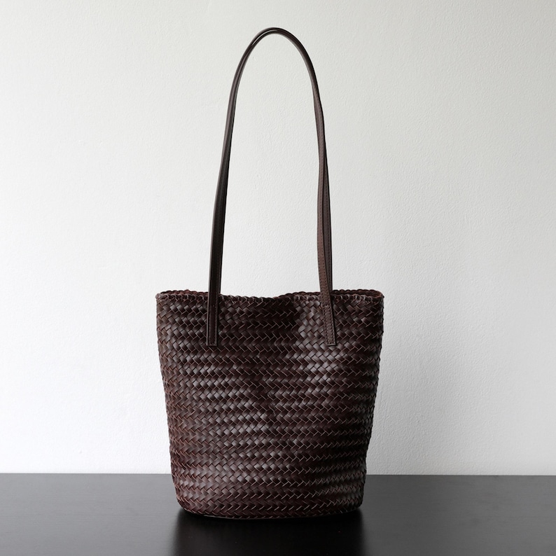 Peut inclure: Un sac fourre-tout en cuir tress&eacute; marron fonc&eacute; avec de longues et fines anses. Le sac a une forme rectangulaire avec un motif tiss&eacute; sur tout le corps. Les anses sont fix&eacute;es en haut du sac et sont en cuir marron fonc&eacute;.