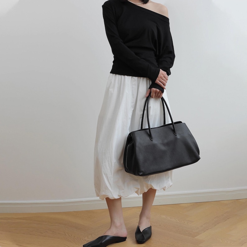 K&ouml;nnte beinhalten: Eine schwarze Tragetasche mit langen Henkeln. Die Tasche wird von einer Person getragen, die ein schwarzes schulterfreies Langarm-Oberteil, einen wei&szlig;en Rock und schwarze Schuhe tr&auml;gt. Die Tasche scheint aus Leder zu sein.