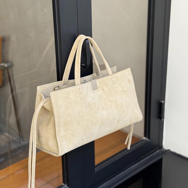 May include: A beige suede tote bag with two handles and a rectangular shape. The bag has a silver-colored clasp and long, thin tassels on the sides. The bag is hanging on a black door frame.