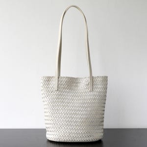 Peut inclure: Un sac fourre-tout blanc tiss&eacute; avec de longues et fines anses. Le sac a un motif en osier et une forme rectangulaire. Les anses sont fix&eacute;es en haut du sac avec de petits morceaux de mati&egrave;re rectangulaires.