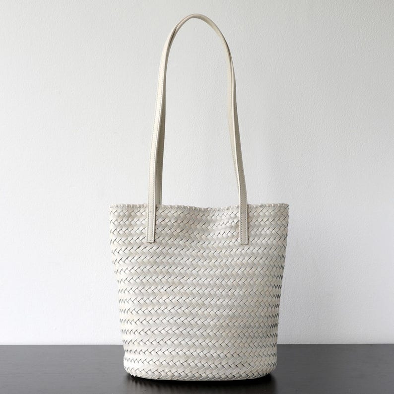 Peut inclure: Un sac fourre-tout blanc tiss&eacute; avec de longues et fines anses. Le sac a un motif en osier et une forme rectangulaire. Les anses sont fix&eacute;es en haut du sac avec de petits morceaux de mati&egrave;re rectangulaires.