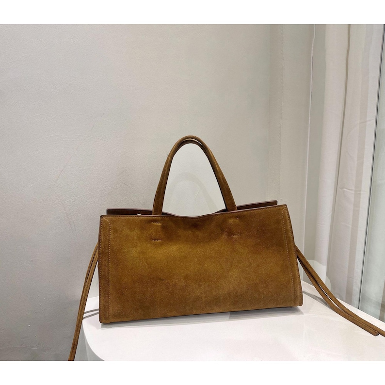 May include: A rectangular brown suede tote bag with two handles and long straps. The bag is sitting on a white surface. The bag has a simple, minimalist design.