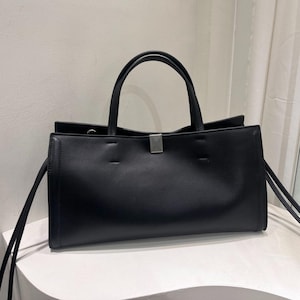 May include: A black tote bag with two handles and a rectangular shape. The bag has a silver-colored clasp and long, thin straps on the sides. The bag is sitting on a white surface.