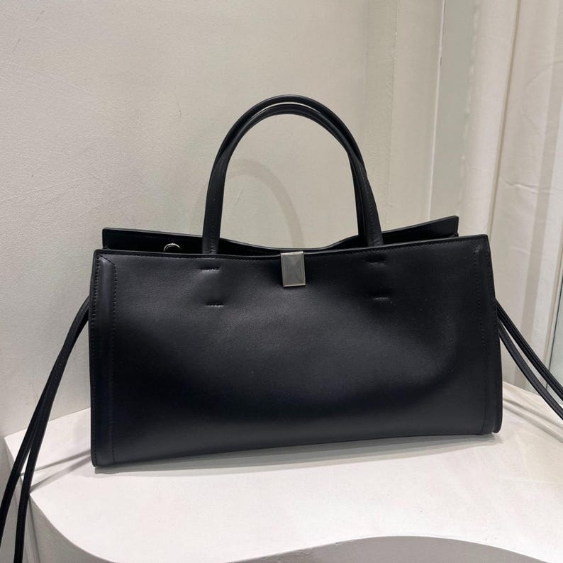 May include: A black tote bag with two handles and a rectangular shape. The bag has a silver-colored clasp and long, thin straps on the sides. The bag is sitting on a white surface.
