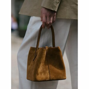 May include: A brown suede handbag with a braided handle. The bag has a structured shape with a slightly flared base. The handle is a darker brown, and the bag appears to be empty. The bag is held by a person wearing a tan jacket and white pants.