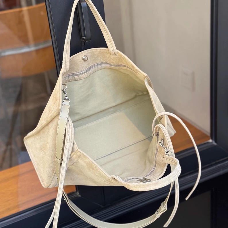 May include: A beige suede tote bag with a zippered top and a detachable shoulder strap. The bag has a spacious interior and a small zippered pocket. The bag is made of a soft, textured material and has a minimalist design.