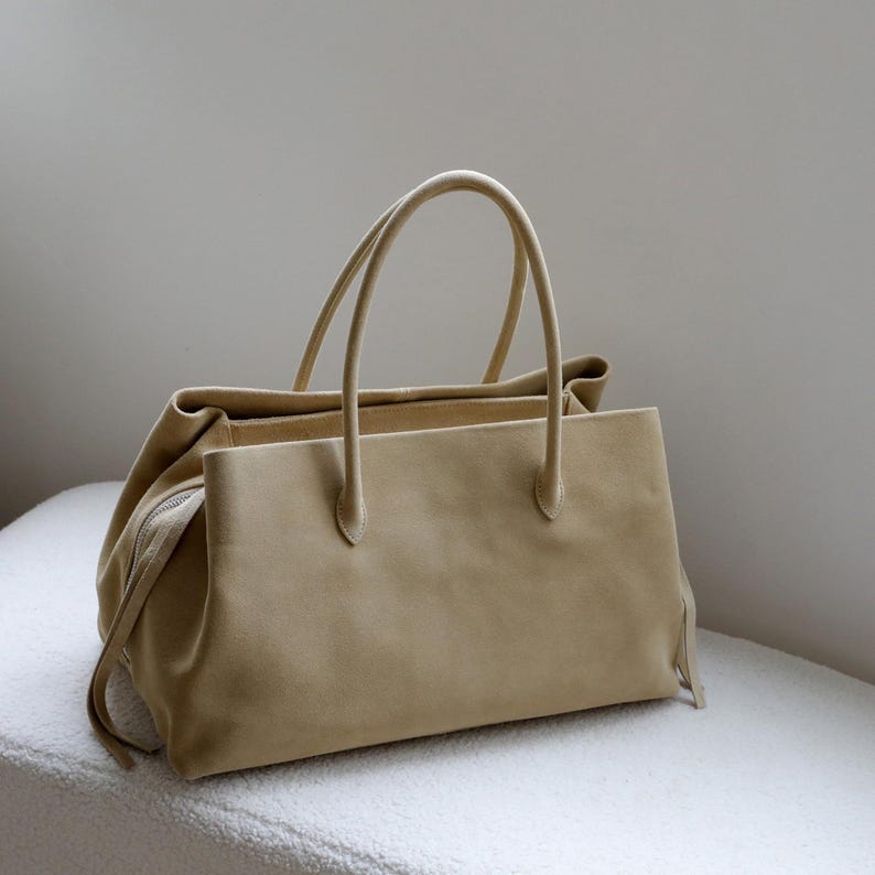 K&ouml;nnte beinhalten: Beige Wildleder-Tote-Tasche mit zwei langen Griffen. Die Tasche hat eine rechteckige Form mit Rei&szlig;verschluss und ein weiches, strukturiertes Aussehen. Die Tasche steht auf einer wei&szlig;en Oberfl&auml;che.