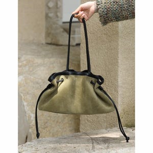 Puede incluir: Un bolso de mano de ante verde oliva suave con un borde de cuero negro en la parte superior y dos correas largas de cuero negro. El bolso cuenta con un cierre de cordón con finas tiras de cuero negro que cuelgan a ambos lados de la estructura.