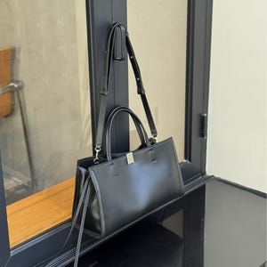 May include: Black rectangular handbag with a top handle and a detachable shoulder strap. The bag has a silver-colored clasp and is hanging near a window.