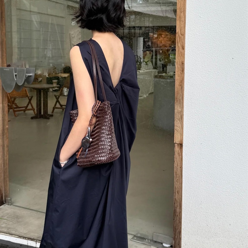 Peut inclure: Une robe sans manches bleu marine fonc&eacute; avec un dos nu. Un sac fourre-tout en cuir tress&eacute; marron avec une bandouli&egrave;re est port&eacute; sur le bras. Le sac a un porte-cl&eacute;s attach&eacute;. L'arri&egrave;re-plan montre une porte vitr&eacute;e et un aper&ccedil;u d'un caf&eacute;.