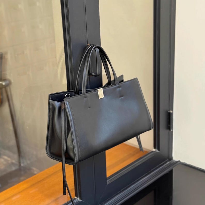 May include: A black leather tote bag with two handles and a rectangular shape. The bag has a gold-colored metal clasp and long, thin tassels on the sides. The bag is hanging on a black door frame.