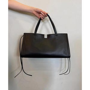 May include: A black tote bag with a rectangular shape and a silver-colored metal accent. The bag has two handles and long, thin straps hanging from the sides. The bag is held up by a hand.