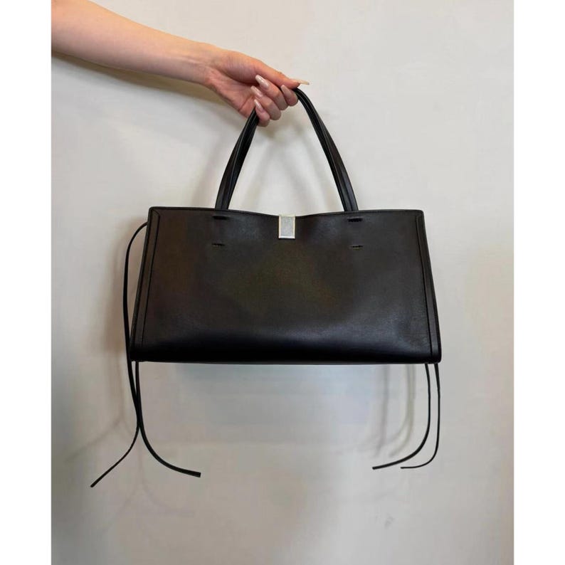 May include: A black tote bag with a rectangular shape and a silver-colored metal accent. The bag has two handles and long, thin straps hanging from the sides. The bag is held up by a hand.